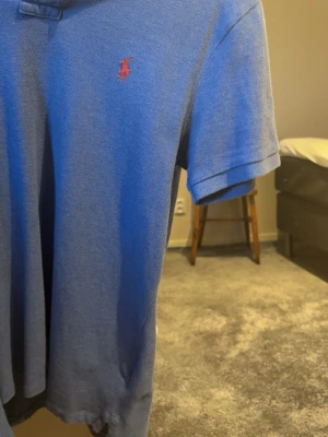 Blå pikétröja från Ralph Lauren - Snygg blå pikétröja från Ralph Lauren med röd broderad logga på bröstet. Modellen är custom slim fit och har korta ärmar samt klassisk krage. Perfekt för en stilren och avslappnad look.