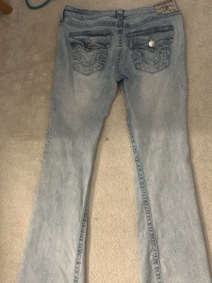 Ljusblå bootcut jeans från True Religion - Säljer ett par ljusblå bootcut jeans från True Religion med klassiska broderade bakfickor och silverfärgade knappar. 