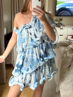 Blåvit oneshoulder volangklänning - Supersöt kort klänning i blått och vitt med blommigt mönster. Klänningen har oneshoulder-design med volanger. Den är ifrån NA-KD och är helt slutsåld. Köpte den på Plick, men ändrade mig att jag ville ha en annan klänning. Går troligtvis ner i pris vid snabb affär, andvänd gärna ”köp nu”