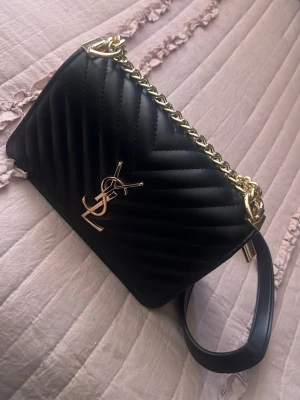 Svart YSL handväska i läder - Säljer en svart quiltad handväska från Saint Laurent med guldfärgade detaljer och kedjerem. Väskan har det klassiska YSL-emblemet framtill och är tillverkad i slätt läder med elegant V-mönster. Perfekt accessoar för dig som gillar lyxig och tidlös design.