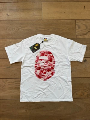 BAPE (A Bathing Ape) - Vit t-shirt från BAPE med det ikoniska apansikte-motivet i röd, rosa och vit camo på både fram- och baksidan. Klassisk passform och rund hals. Perfekt statement-plagg för streetwear-stilen.
