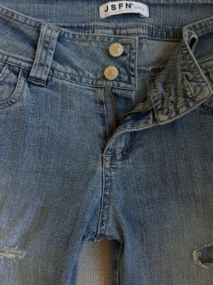 Ljusblå slitna jeans från JSFN - Säljer ett par ljusblå jeans från JSFN med slitningar på benen och två knappar i midjan. Lågmidjade och bootcut.
