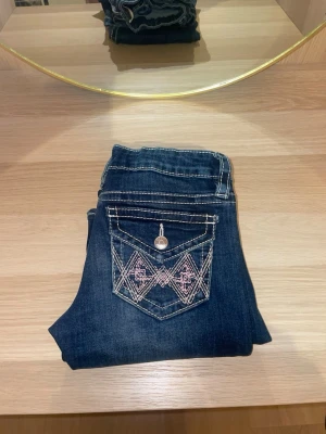 Mörkblå bootcut jeans med broderad ficka från ginatricot  - Säljer ett par mörkblå bootcut jeans med snyggt broderad bakficka i vitt och rosa i storlek 152. Jeansen har låg midja och kontrastsömmar som ger en cool look. Perfekta för dig som gillar detaljer och vill sticka ut lite extra. Skriv för fler bilder, pris kan diskuteras!💗