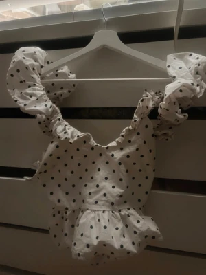 Vit prickig blus med volanger - Supersöt vit blus med svarta prickar och stora volangärmar. Djup rygg med knytband och volangdetaljer även i midjan. Perfekt för dig som gillar en lekfull och trendig stil. Tunn och luftig bomullskänsla.
