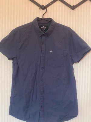 Blå kortärmad skjorta från Hollister - Säljer en blå kortärmad skjorta från Hollister med klassisk krage och knappar hela vägen fram. Skjortan har en liten vit logotyp broderad på bröstet och är tillverkad i bomull. Perfekt för dig som vill ha en enkel och stilren look.
