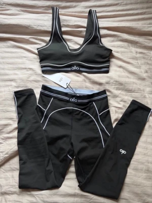 Svart träningsset från Alo - Snyggt svart träningsset från Alo med sport-bh och tights. Setet har vita kontrastsömmar och logga på både bh och byxa. Materialet är stretchigt och följsamt, perfekt för träning eller chill. Sport-bhn har bred resår och tightsen har hög midja för extra stöd.