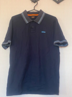Marinblå pikétröja från BOSS - Snygg marinblå pikétröja från BOSS med klassisk krage och två knappar framtill. Tröjan har korta ärmar med kontrasterande ribbade muddar och BOSS-logga broderad på bröstet. Perfekt till en avslappnad men stilren look.