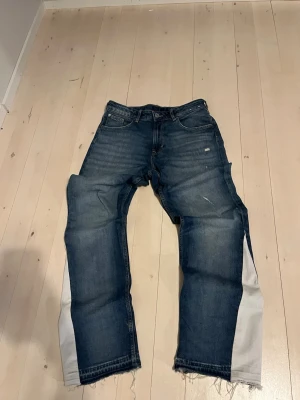 Blå jeans med vit panel och råa kanter - Unika blå jeans med vit panel längs utsidan av benen och råa, fransiga benslut. Jeansen har en klassisk femficksmodell och subtila slitningar framtill för en trendig look. Perfekta för dig som vill sticka ut med en cool och avslappnad stil.