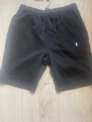 Svarta shorts från Polo Ralph Lauren - Svarta shorts från Polo Ralph Lauren med klassisk vit logga broderad på vänster ben. Shortsen har snörning i midjan och är tillverkade i mjuk bomull, perfekta för en avslappnad stil. Två sidofickor och bekväm passform.