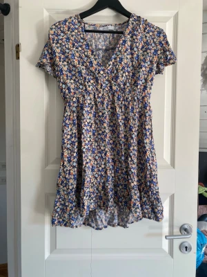 Blommig kortklänning från Amisu - Säljer en söt kortklänning från Amisu med blommigt mönster i blått, beige, orange och vitt. Klänningen har v-ringad hals, korta ärmar och volangdetalj nedtill. Materialet är lätt och känns som polyester, perfekt för varma dagar.