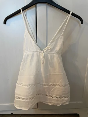 Vit linnetopp med spetsdetaljer - Söt vit linnetopp med tunna axelband och v-ringning. Toppen har knyt framtill och dekorativa spetsdetaljer längs nederdelen. Toppen är använd 1 gång och säljs då jag har en i storleken större som passar bättre. Köpt från Gina tricot och säljs för 150kr men pris kan diskuteras. OBS! Köparen står för frakt. 