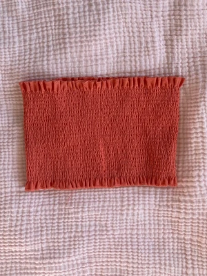 Rostfärgad bandeau tubtopp - Säljer en rostfärgad bandeau tubtopp med smockad struktur och elastiska kanter upptill och nedtill. Toppen är axelbandslös och har en tight passform som sitter snyggt mot kroppen. Perfekt för varma dagar och sommarhäng.