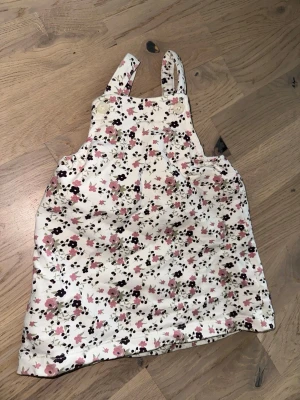 Blommig kort klänning - Blommig kort klänning Supersöt kort klänning  och blommigt mönster i rosa, svart och beige på vit botten. Klänningen har knappar vid axelbanden och är perfekt för en avslappnad och lekfull stil.