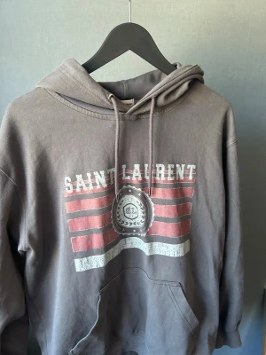 Grå Saint Laurent hoodie med tryck - Säljer en grå hoodie från Saint Laurent med vitt och rött tryck framtill och texten 'SAINT LAURENT'. Hoodien har huva med dragsko och en stor magficka. Tillverkad i mjukt bomullsmaterial och har en avslappnad passform, perfekt för en chill och trendig look.