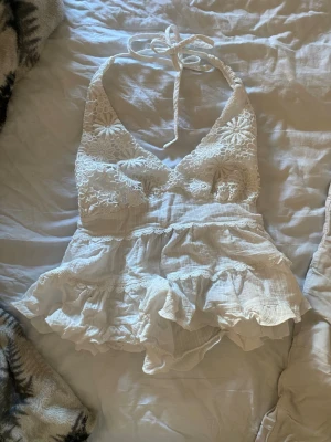 Vit spetslinnetopp med volanger - Söt vit linnetopp med spets och broderade blommor upptill. Toppen har halterneck med knyt i nacken, v-ringning och volangkant nedtill. Tillverkad i bomull och linne för en luftig känsla, perfekt för sommaren.
