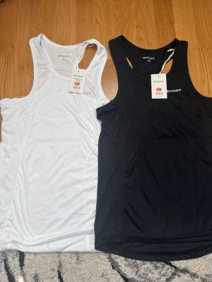 Träningslinnen - Två träningslinnen från SPORT i storlek M, ett svart och ett vitt. Båda är tillverkade i ett lätt och snabbtorkande funktionsmaterial, perfekta för gymmet eller löprundan. Klassisk racerback-modell för maximal rörelsefrihet.