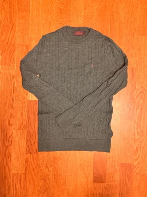 Grön Morris tröja - Snygg grå kabelstickad tröja från Lexington med rund halsringning och diskret rosa broderad logga på bröstet. Tröjan har långa ärmar och ribbade muddar vid ärmslut och nederkant. Perfekt för dig som gillar klassisk och stilren design.