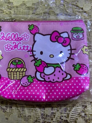 Hello Kitty rosa plånbok - Söt rosa plånbok med Hello Kitty-motiv och vita prickar. Plånboken har färgglada detaljer med jordgubbar, stjärnor och texten 'Hello Kitty'. Tillverkad i slitstarkt tygmaterial och stängs med dragkedja. Perfekt för småsaker och kort. 49kr/st 