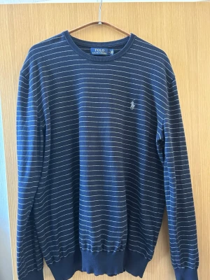 Marinblå randig tröja Polo Ralph Lauren - Snygg marinblå tröja från Polo Ralph Lauren med tunna vita ränder och klassisk logga broderad på bröstet. Tröjan har rund halsringning, långa ärmar och ribbade muddar vid ärmslut och nederkant. Perfekt för en stilren och avslappnad look.