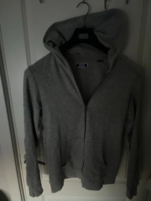 Grå zip hoodie från Jack & Jones - Snygg grå hoodie från Jack & Jones med dragkedja framtill och två stora fickor. Tillverkad i mjukt bomullsmaterial och har en klassisk huva. Perfekt för en avslappnad stil och enkel att matcha med andra plagg.