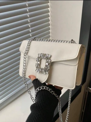 Vit kuvertväska med strassdetalj - Snygg vit kuvertväska i rektangulär form med silverfärgad kedjerem och stor glittrig strassdetalj på locket. Väskan har en stilren design och passar perfekt till att lyfta vilken outfit som helst. Materialet ser ut att vara konstläder.