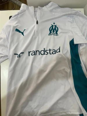 Marseille träningströja Puma vit - Säljer en vit långärmad träningströja från Olympique de Marseille med turkosa detaljer och halv dragkedja. Puma-logga på bröstet, Marseille-klubbmärke och sponsortryck från Randstad. Tillverkad i lätt och ventilerande funktionsmaterial.