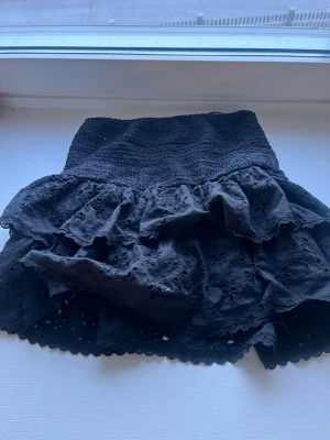 Svart volangkjol från Gina Tricot - Kort svart kjol från Gina Tricot med bred smock i midjan och dubbla volanger. Kjolen har broderade detaljer och hålmönster som ger en romantisk vibe. Perfekt att matcha med en enkel topp för en trendig look.