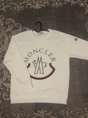 Vit Moncler hoodie med stor logga - Säljer en vit hoodie från Moncler med stor ikonisk logga i svart, rött och blått på bröstet. Storlek S men passar bra med M