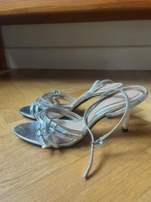 Silvriga glittriga pumps med remmar - Säljer ett par silvriga klackar, smala remmar och öppen tå. Skorna har en silvrig finish, liten klack och justerbart spänne runt ankeln. 