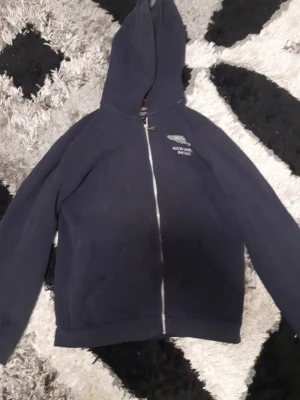 Mörkblå Aston Martin Racing hoodie - Snygg mörkblå hoodie med dragkedja från Aston Martin Racing. Broderad logga och text på bröstet. Klassisk huva och två fickor framtill. Perfekt för dig som gillar motorsport och vill ha en stilren look. ( size L på barn size )