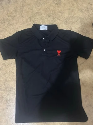 Svart piké från Ami med rött hjärta - Säljer en nästan helt ny och oanvänd S/XS svart pikétröja från Ami med klassisk krage och knappar framtill. På bröstet finns ett broderat rött hjärta med bokstaven A. Tillverkad i bomull för en bekväm känsla och stilren look.
