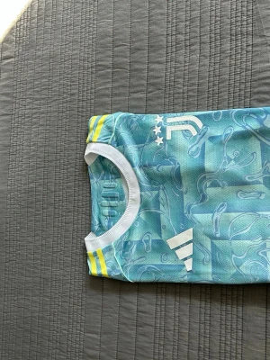 Juventus tränings t-shirt Adidas blå - Juventus tränings t-shirt från Adidas i ljusblått med vitt och gula detaljer. Tröjan har ett abstrakt mönster, vita ränder på axlarna och Juventus-logga med tre stjärnor på bröstet. Tillverkad i lätt och ventilerande material, perfekt för träning.