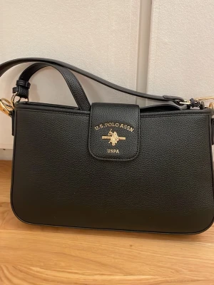 Svart handväska från U.S. Polo Assn. - Stilren svart handväska från U.S. Polo Assn. med guldfärgade detaljer och logotyp framtill. Väskan har en rektangulär form, justerbar axelrem och är tillverkad i ett slätt, konstläderliknande material. Perfekt för dig som gillar klassisk och enkel design.
