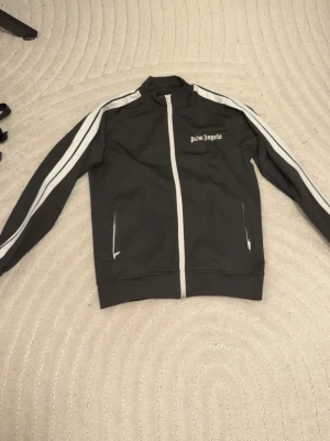 Svart track jacket från Palm Angels - Snygg svart track jacket från Palm Angels med vita ränder längs ärmarna och vit dragkedja. Jackan har hög krage, två fickor med dragkedja och logga på bröstet. Perfekt för en sportig och trendig look.