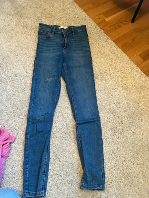 Blå tighta jeans med hög midja från gina tricot - Säljer ett par klassiska blå jeans med hög midja och smal passform. Jeansen har fem fickor och dragkedja med knapp framtill. Perfekta till vardagslooken och passar till det mesta. Materialet är mjukt och stretchigt för extra komfort.