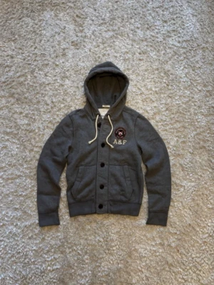 Grå hoodie från Abercrombie & Fitch strl S - Snygg grå hoodie med knappar framtill från Abercrombie & Fitch. Har broderad logga och emblem på bröstet, stora fickor och vita dragsnören i huvan. Tillverkad i mjukt sweatshirtmaterial, perfekt för en avslappnad stil. Tröjan är väldigt svår att få tag på i bra skick. Passar ~ 170-180 cm. Hör av er vid frågor och funderingar!