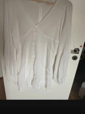 Vit omlottklänning med knappar - Säljer en vit kortklänning med omlottdesign och v-ringning. Klänningen har långa ärmar, knappar framtill och lätt struktur i tyget. Perfekt för dig som gillar en enkel men stilren look.