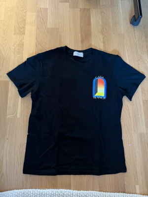 Svart Casa Blanca t-shirt med tryck - Säljer en svart t-shirt från Casa Blanca med färgglatt grafiskt tryck fram och stort tryck bak i blått, gult, rött och vitt. Klassisk passform och rund hals. Perfekt för dig som gillar streetwear och vill sticka ut med snygg design.