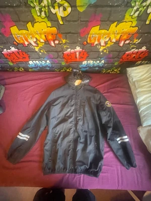 Svart vindjacka med reflexdetaljer - Svart vindjacka från Vettelli med huva och dragkedja framtill. Jackan har reflexdetaljer på båda ärmarna och ett runt tygmärke på vänster ärm. Två sidofickor med dragkedja och resår i nederkant och ärmslut. Perfekt för blåsiga dagar.