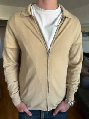 Beige ziptröja från J.Lindeberg - Snygg beige ziptröja från J.Lindeberg i mjukt material. Tröjan har hel dragkedja framtill, ribbade muddar och klassisk krage. Perfekt att slänga över en t-shirt för en clean och stilren look.