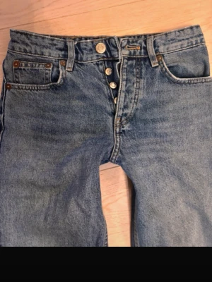 Blå raka jeans med knappar - Säljer ett par klassiska blå jeans med raka ben och hög midja. Jeansen har knappgylf, fem fickor och råa kanter vid bensluten. Perfekta för en avslappnad och trendig look. Materialet är kraftig denim i bomull.
