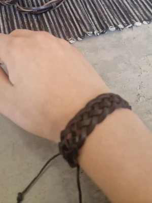 Flätat brunt läderarmband - Snyggt flätat armband i mörkbrunt läder med justerbar knytning. Armbandet har en bred och robust design som ger en cool och avslappnad vibe. Perfekt accessoar för dig som gillar en enkel men stilren look.