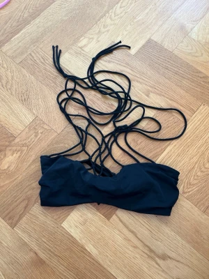 Svart bikiniöverdel med snörning - Trendig svart bikiniöverdel med långa tunna snören som kan knytas på olika sätt för en personlig look. Bandeau-modell utan vaddering och utan mönster. Perfekt för sommardagar på stranden eller vid poolen.