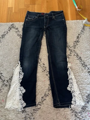 Lågmidjade mörkblå jeans med spetsdetaljer - Unika mörkblå jeans med utsvängda ben och vita virkade spetsdetaljer längs bensluten. Klassisk femficksmodell med kontrastsömmar och låg midja. Perfekta för dig som vill sticka ut med en kreativ och bohemisk stil.