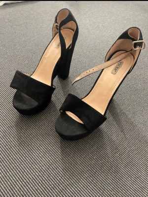 Svarta platåpumps från Vices - Säljer ett par svarta pumps från Vices med bred klack och platåsula. Skorna har öppen tå, justerbar rem med guldfärgat spänne och är klädda i mockaimitation. Perfekta för dig som gillar chunky klackar och stilren design.