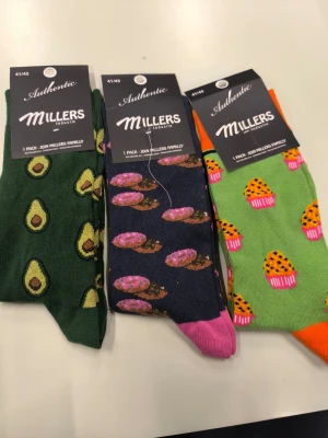 Färgglada strumpor med mönster Millers - Tre par strumpor från Millers med olika mönster: gröna med avokado, mörkblå med donuts och ljusgröna med muffins. Strumporna har färgglada detaljer vid tå och häl och är tillverkade i mjukt material. Perfekta för dig som gillar roliga och unika accessoarer.
