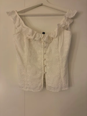 Vit blommig blus med volang - Söt vit blus med broderade blommor, volangkant vid axlarna och knappar framtill. Ärmlös modell. 