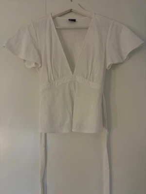 Vit topp med v-ringning med knyt på baksidan  - Säljer en vit topp med v-ringning och korta, vida ärmar. Toppen har knytband i ryggen. 