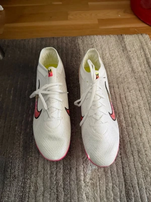 Nike Mercurial Vita Fotbollsskor - Säljer ett par Nike Mercurial fotbollsskor i vitt med rosa och svarta detaljer. Skorna har en lätt ovandel i syntetmaterial och en sula med rosa och iriserande dobbar för grymt grepp på planen. Swoosh-logga på sidan och snörning för tajt passform.