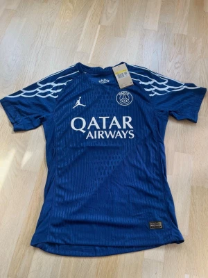 PSG fotbollströja - Snygg blå PSG matchtröja med vita detaljer och Qatar Airways-tryck på bröstet. Tröjan har korta ärmar, rund hals och coola mönster på axlarna. Tillverkad i lätt och ventilerande material, perfekt för träning eller match.